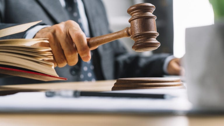 This is a picture of a man's torso and arms, shown in a business suit in the process of banging a legal judge's gavel onto its base, with an open text book next to him.